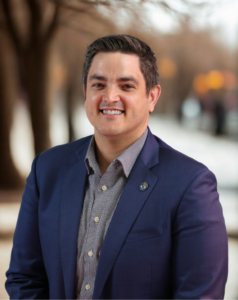 Nicholas Kiego in a dark blue blazer and button-up shirt is smiling with a blurred background