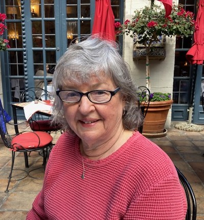 An older woman with gray hair and glasses is sitting outdoors at a cafe with red umbrellas and potted plants, wearing a red sweater.
