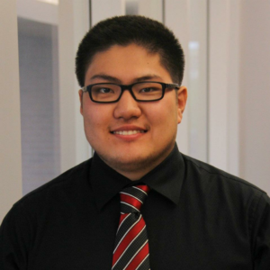 A person wearing glasses, a black shirt, and a striped red and black tie smiles at the camera.