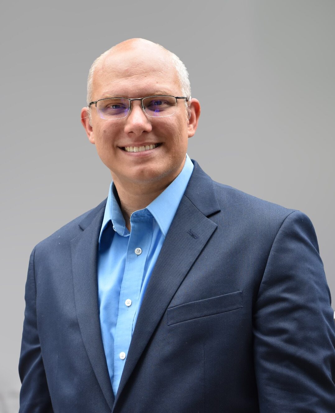 A man wearing glasses, a blue button-down shirt, and a dark blue suit jacket smiles at the camera against a gray background.